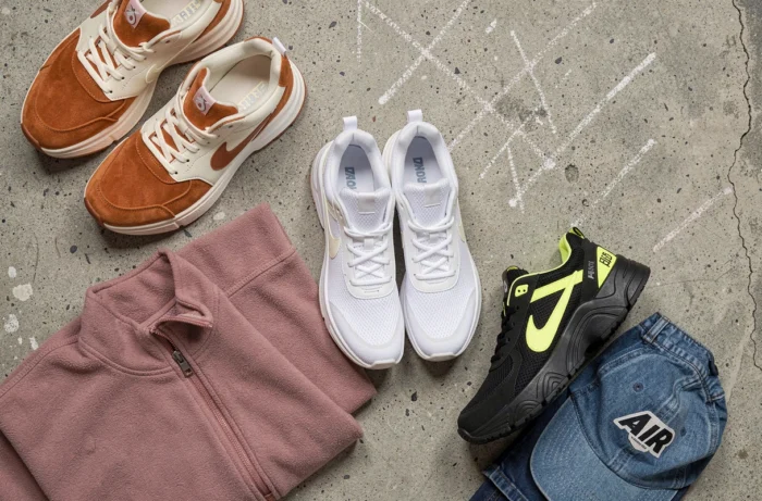 Flat-lay de trois paires de sneakers tendance en suède, mesh et plateforme chunky sur béton brut, entourées de pièces streetwear pliées dont un zip polaire rose et une casquette denim